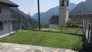 A stone-paved courtyard with a grassy area, a white building with a bell tower, and mountains in the background.