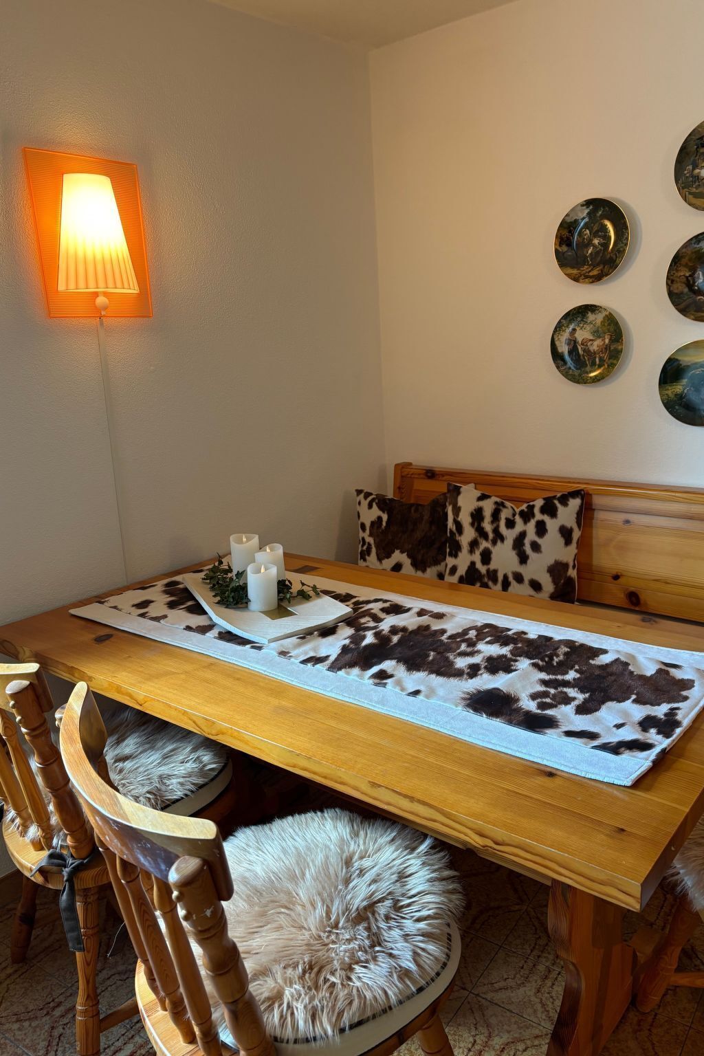 A dining area with a wooden table, chairs, a cowhide placemat, a wall-mounted lamp, and decorative plates.