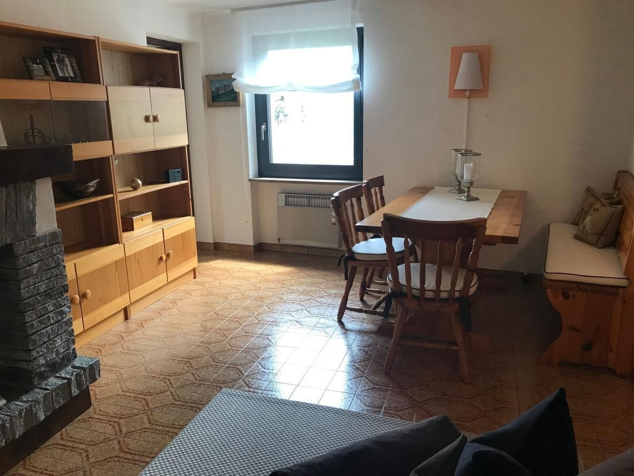 A cozy room with a wooden dining table, chairs, and a bench. There is a window with a curtain, a radiator, and a lamp on the wall.