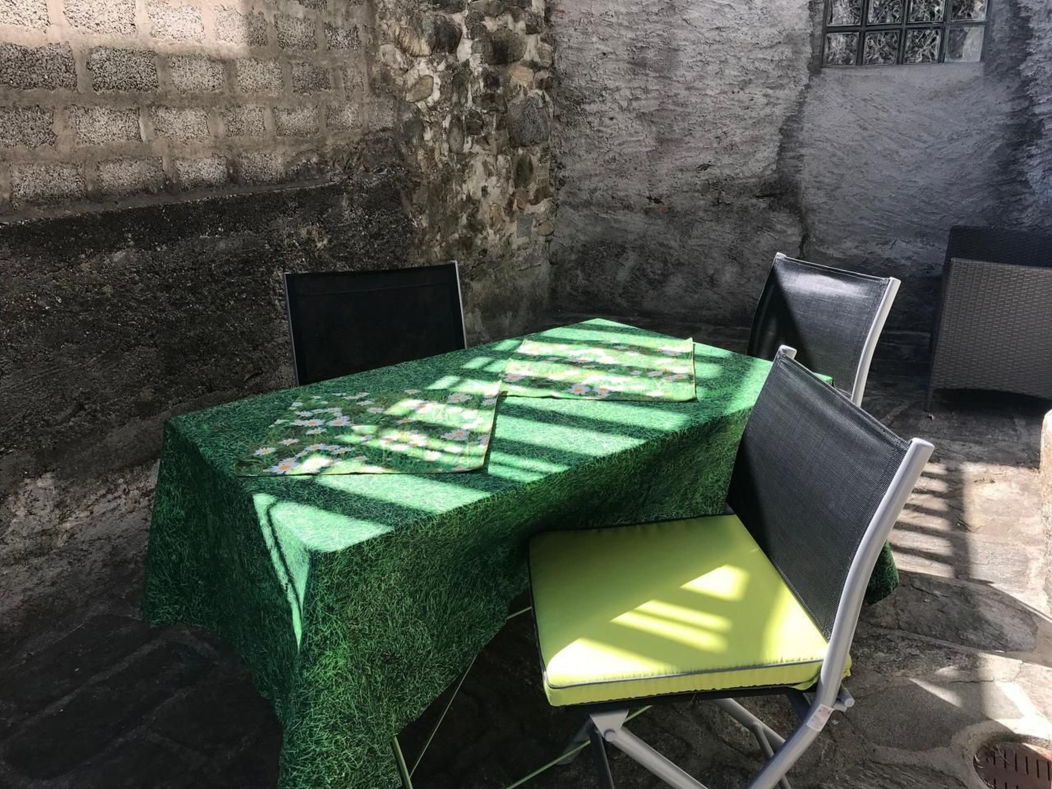 Taula amb estovell verd, dues cadires i una finestra amb vista a una paret de pedra.