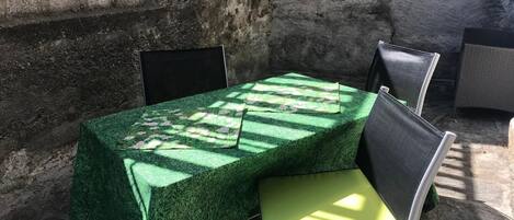 A table with a green tablecloth, two chairs, and a window with a view of a stone wall.