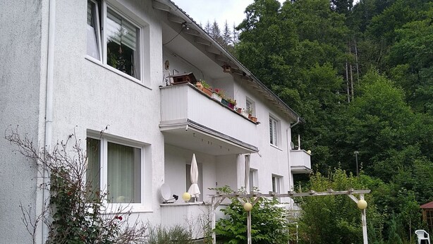 En vit byggnad i två våningar med balkonger, omgiven av grönska och ett trästaket.