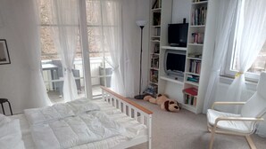 Ett sovrum med en vit säng, en trästol, en bokhylla och ett fönster med tunna, genomskinliga gardiner.