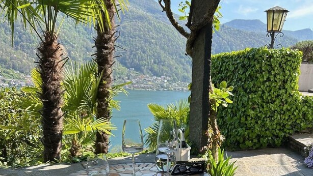 A round table with a mosaic top, two wine glasses, and a bottle, set outdoors with a view of a lake and mountains in the background. - La Quercia by Interhome (Vico Morcote)