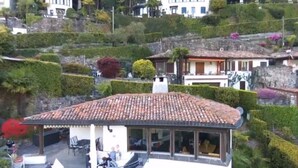 A white building with a tiled roof, a balcony with outdoor seating, and a view of a hillside with other buildings and greenery. - La Quercia by Interhome (Vico Morcote)