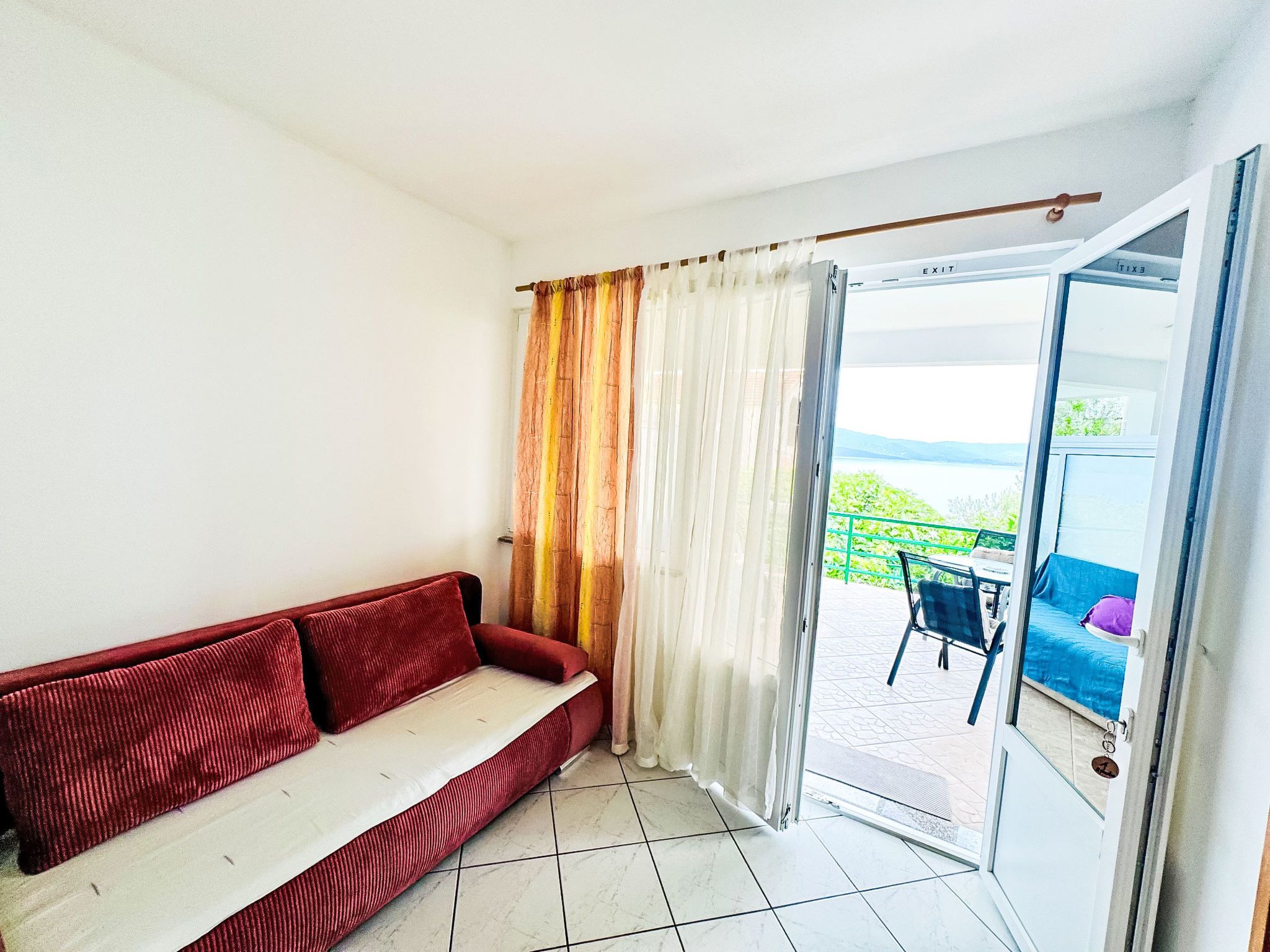 A room with a red sofa, a curtain, and a view of a balcony with outdoor furniture.