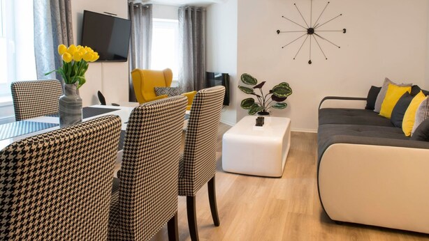 En moderne stue med en sofa, ternede lænestole, et spisebord med blomster i en vase, et fjernsyn og et vægur.
