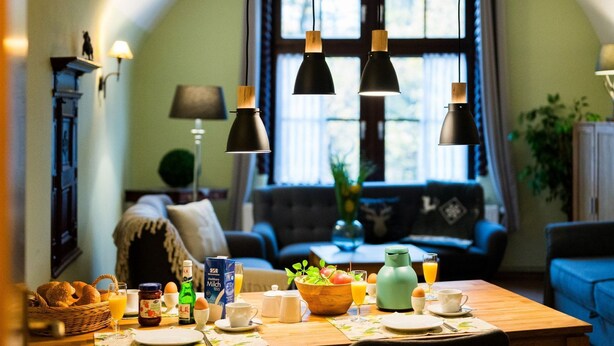 En matplats med ett träbord dukat för frukost, en grön vattenkokare och en korg med bröd.
