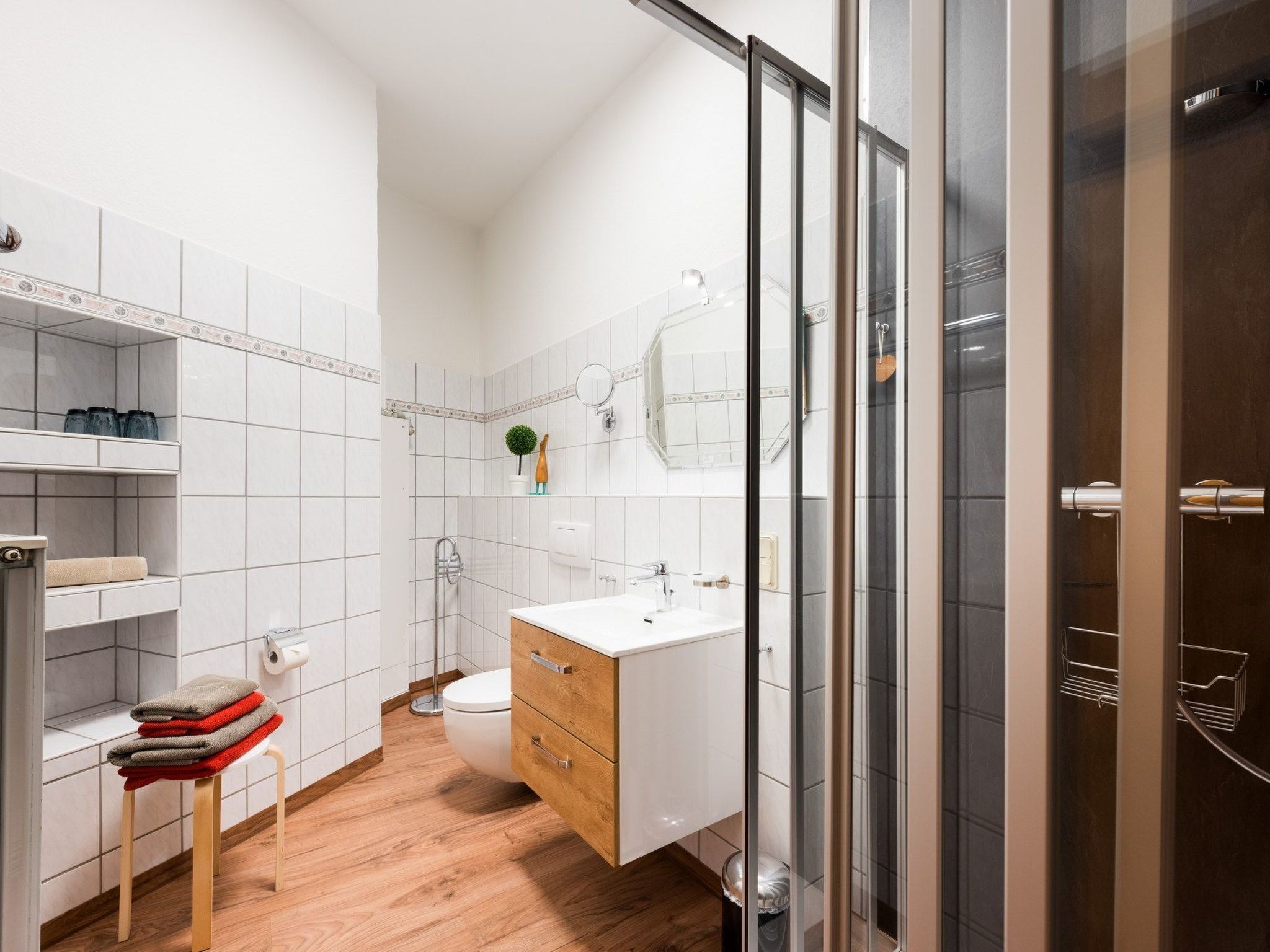 A modern bathroom with white tiles, a wooden floor, a glass shower enclosure, and a white sink with a wooden cabinet.