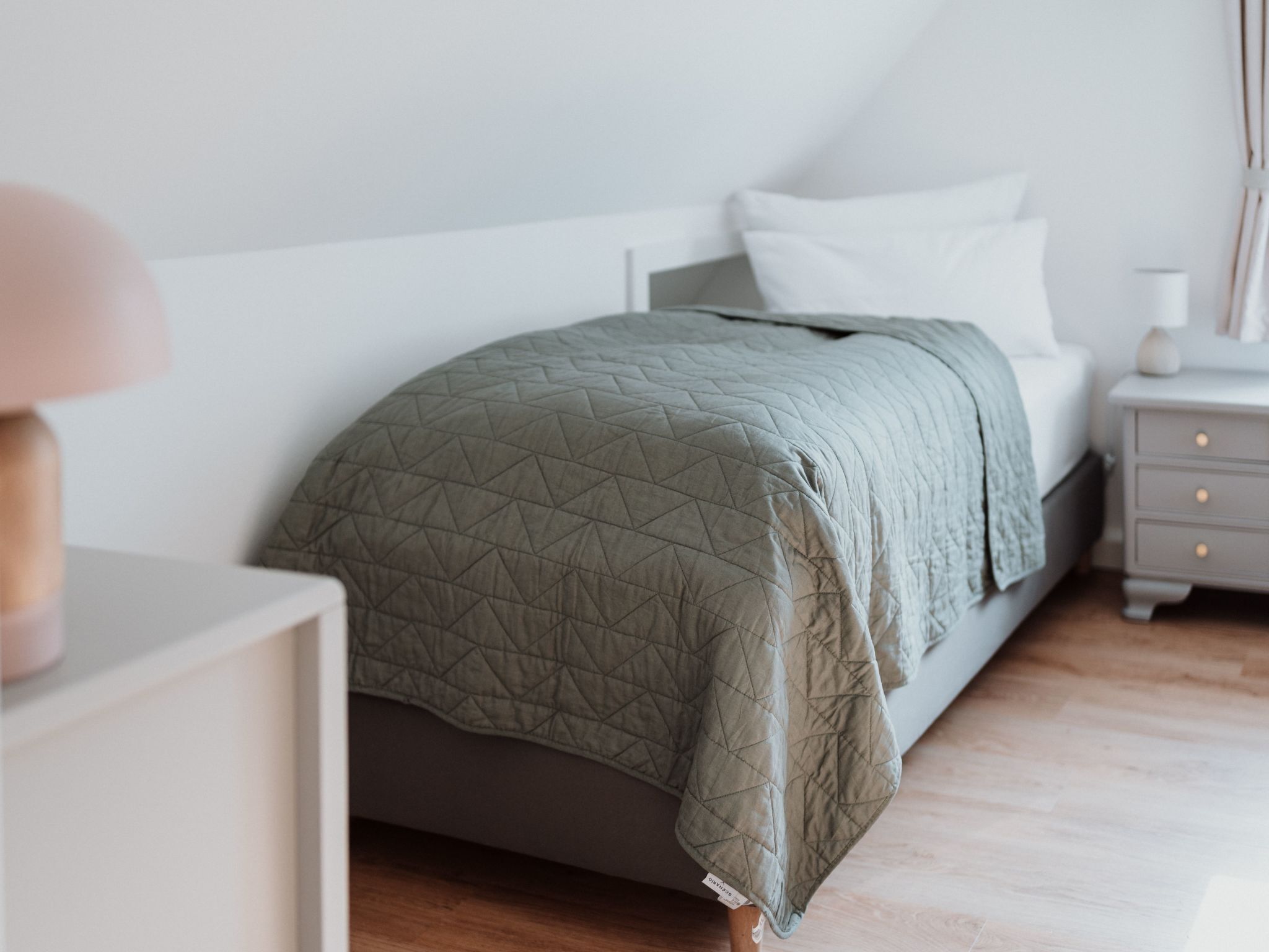 Un letto rifatto con una coperta verde, un comodino con una lampada e un tavolino con cassetti.