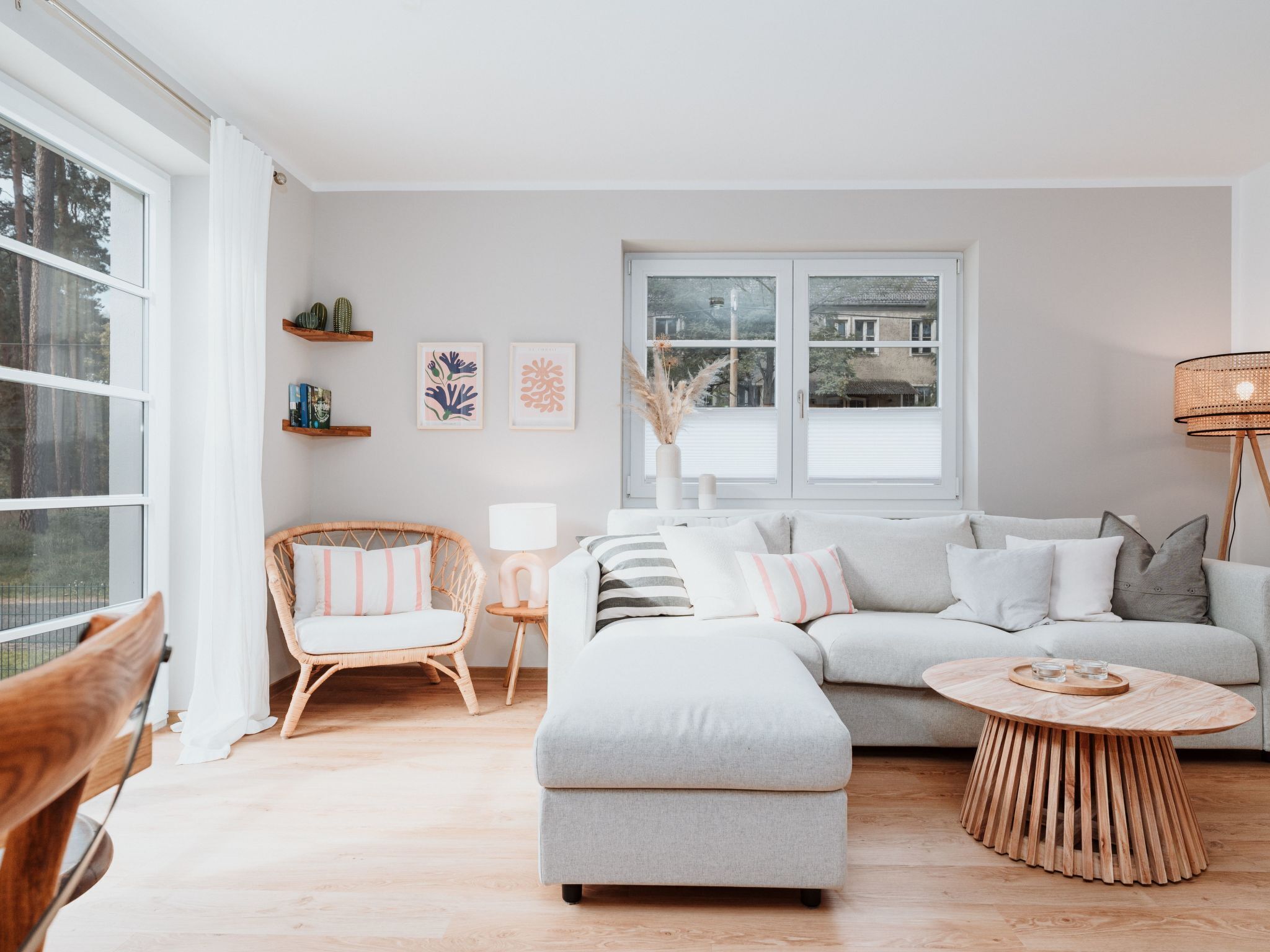 Un salon moderne avec un canapé gris, une table basse en bois et un fauteuil en osier. Il y a deux fenêtres avec des rideaux blancs, une étagère murale avec des livres et des œuvres d’art encadrées.