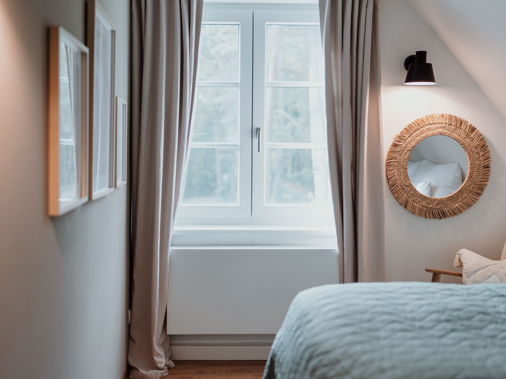 A bedroom with a large window, a bed with a blue blanket, a round mirror, and a wall-mounted light.