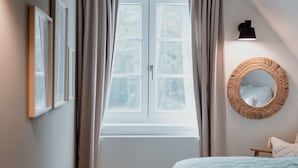 Un dormitorio con un ventanal grande, una cama con una colcha azul, un espejo redondo y una lámpara fijada a la pared.