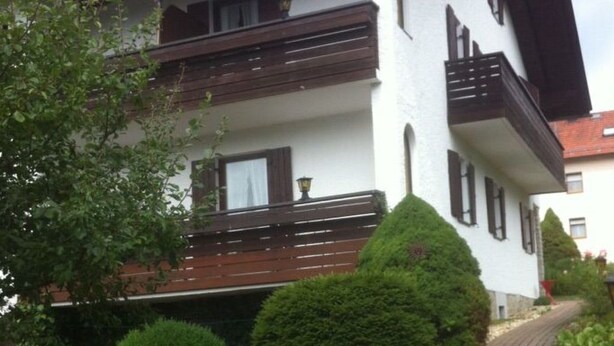 Ein zweistöckiges Gebäude mit weißer Fassade und dunklem Dach. Es besitzt einen Balkon im zweiten Stock sowie einen gepflasterten Weg zum Eingang.