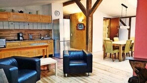 Een moderne keuken met houten kasten, een koelkast en een magnetron. Er is een eethoek met een tafel en stoelen, en een woonkamer met een bank en een salontafel.