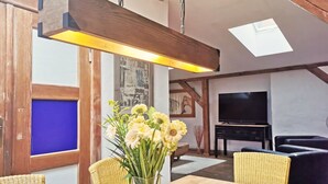 Een eetgedeelte met een tafel, stoelen, een bloevas, een hanglamp en een televisie op de achtergrond.
