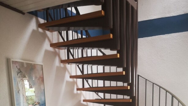 Una scala a chiocciola con gradini in legno e ringhiera in metallo, che conduce a un piano superiore. In primo piano, due poltrone decorate, e un quadro incorniciato appeso al muro.