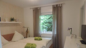 Quarto de hotel com cama, secretária com televisão, janela com cortinas e estante com planta.