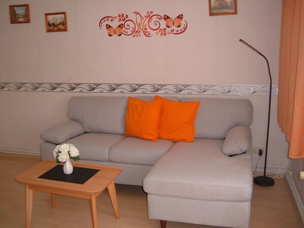 Ein Wohnzimmer mit grauem Sofa, einem orangefarbenen Kissen, einem kleinen Holztisch mit Blumenvase und einer Wanddekoration.