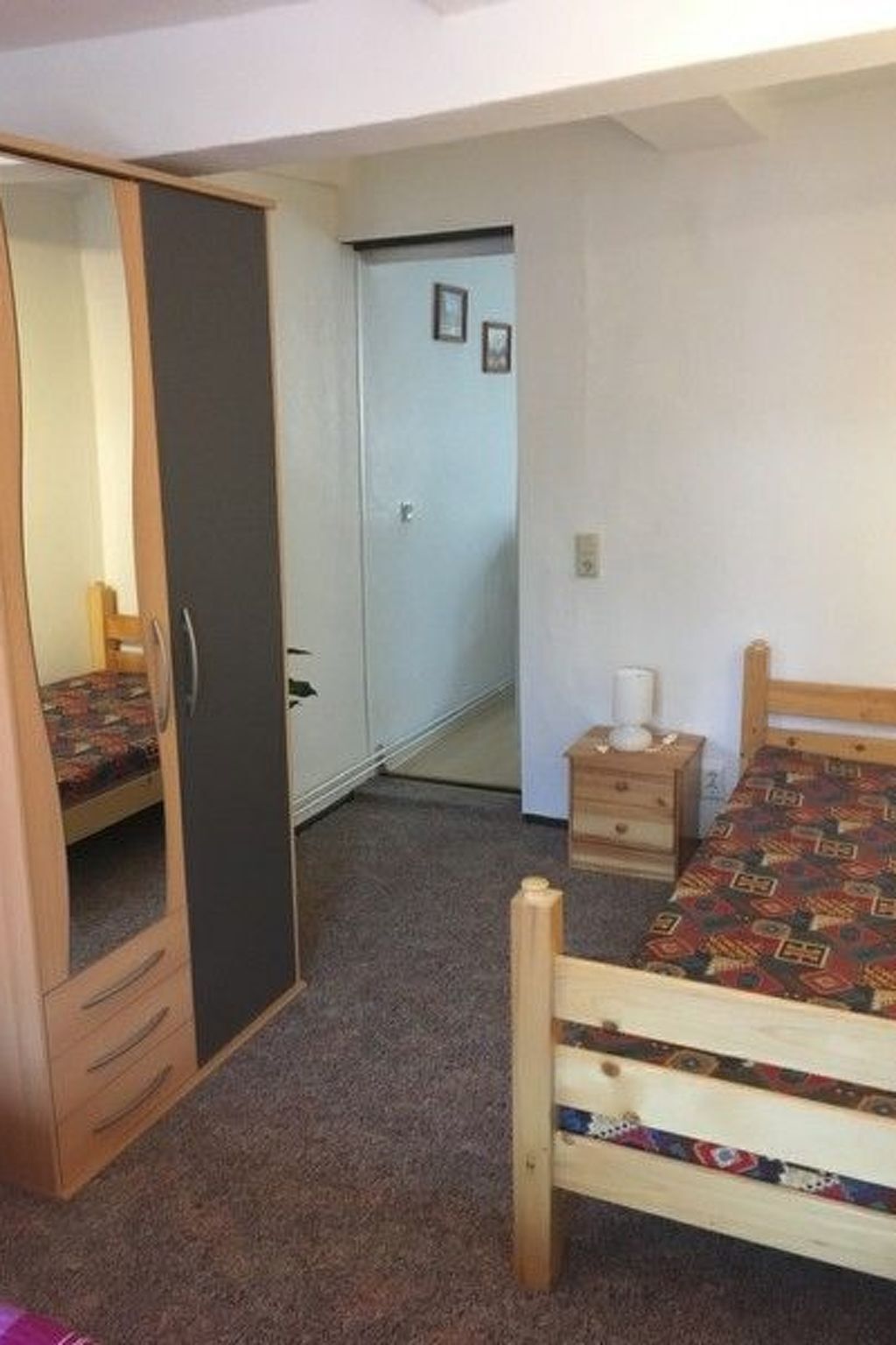 A bedroom with a wooden bed, a wardrobe, a bedside table, and a door leading to another room.