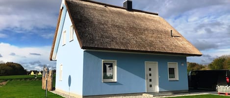 Une maison de plain-pied avec un toit de chaume, une porte blanche et une petite fenêtre à l’avant.