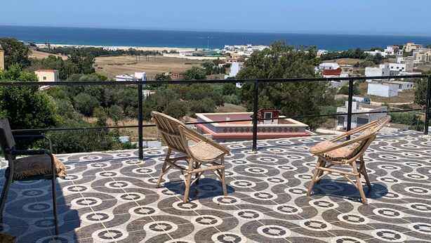 A terrace with patterned flooring, two wicker chairs, and a view of the sea and buildings. - Wohnung Panoramablick by Interhome (Tanger)