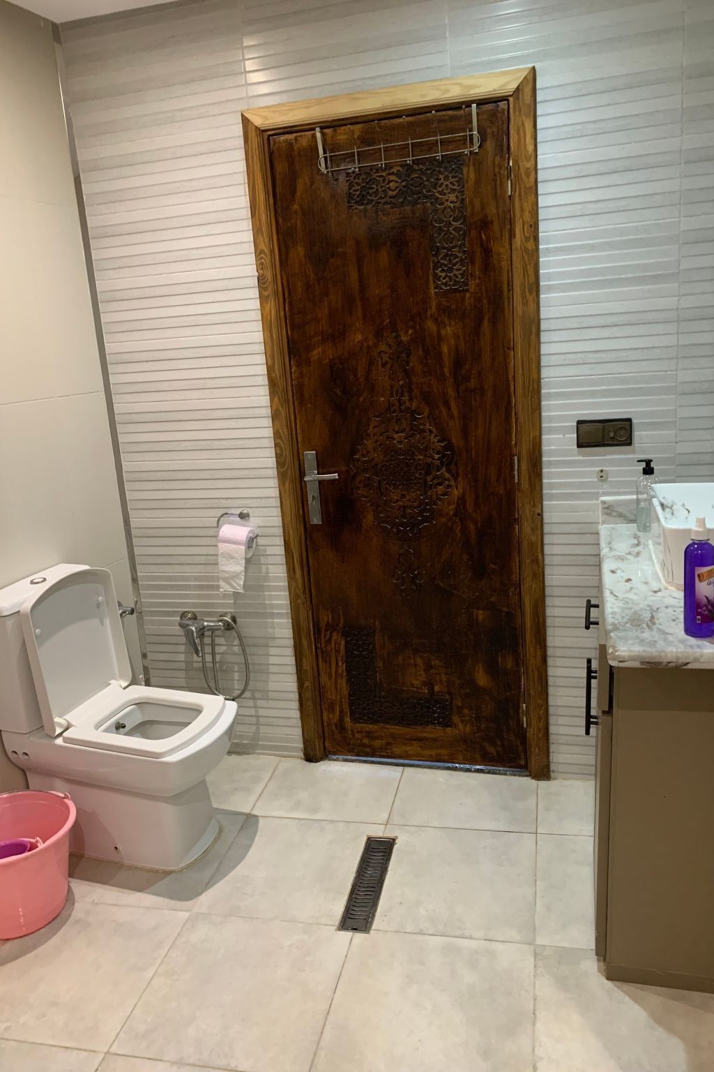 A bathroom with a wooden door, a toilet, a sink with soap dispenser, and a shower.