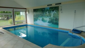 An indoor swimming pool with clear blue water, a tiled floor, and a large window with a view of a garden. - Nette Wohnung mit Garten, Grill und Pool by Interhome (Beetzsee)