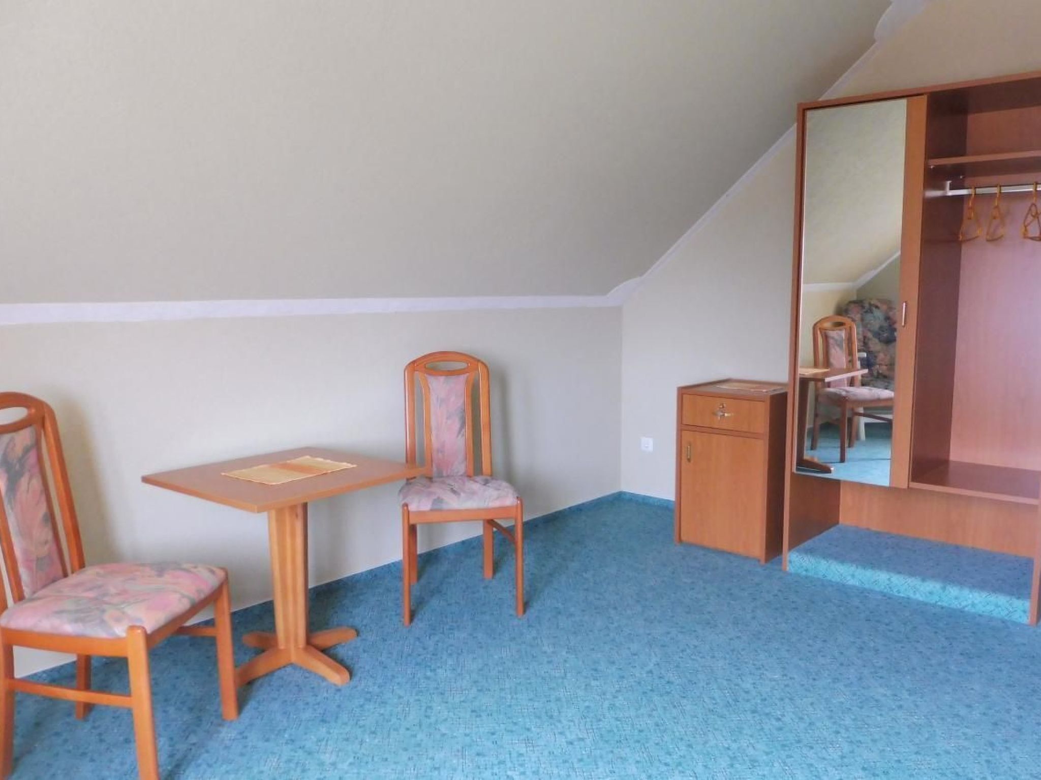 Um quarto pequeno com carpete azul, uma mesa de madeira, duas cadeiras, um armário pequeno e um guarda-roupa com espelho.