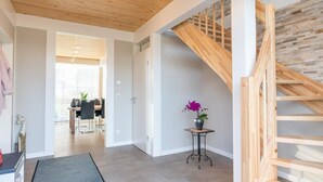 Ein geräumiger Flur mit einer Holztreppe, einem Essbereich im Hintergrund und einem kleinen Tisch mit einer violetten Orchidee.