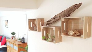 Un salon avec une commode en bois, un canapé avec des coussins rouges et des étagères en bois fixées au mur, ornées d’objets décoratifs.