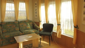 A cozy living room with a patterned sofa, a small table, and a chair. There are large windows with curtains, a framed picture on the wall, and a wooden floor. - mit Pool by Interhome (Beetzsee)