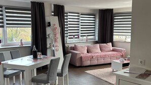 Een moderne woonkamer met een eethoek, met een roze bank, witte meubels en grote ramen met zonwering.