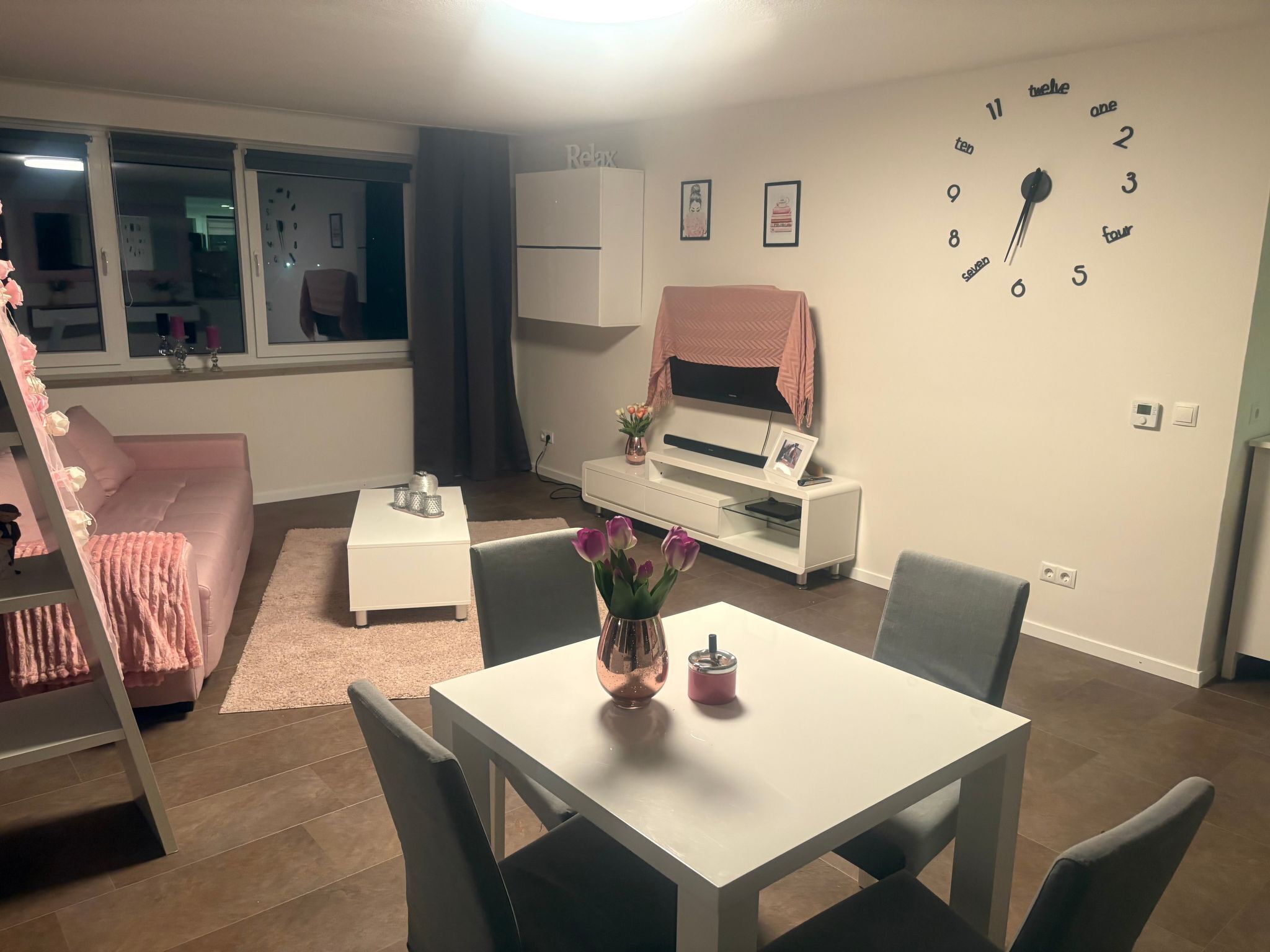 Una sala de estar moderna con un sofá blanco, una mesita pequeña blanca y una mesa de comedor con sillas. Hay un reloj en la pared y un televisor también en la pared.