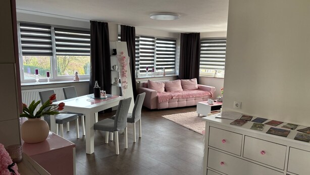 Een moderne woonkamer met een witte eettafel, grijze stoelen, een roze bank en een witte dressoir met lades.