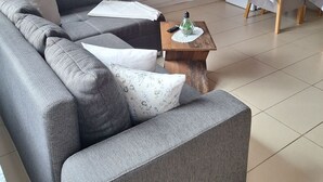 Ein Wohnzimmer mit grauem Sofa, einem kleinen Holztisch mit Fernbedienung, einem an der Wand befestigten Fernseher und einem Fenster mit Blick auf Grünflächen.