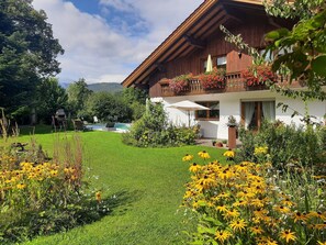 Une maison en bois dotée d’un balcon, d’une piscine et d’un jardin bien entretenu, agrémenté de fleurs jaunes.