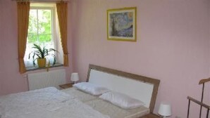 Ein Schlafzimmer mit Bett, Nachttischen, einem Fenster mit Vorhängen, einer Zimmerpflanze und einem Bild an der Wand.