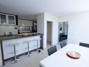 A modern kitchen with a central island, bar stools, and a dining area with a white table and chairs. - 129 on Kloof Nek by Interhome (Cape Town - City Center)