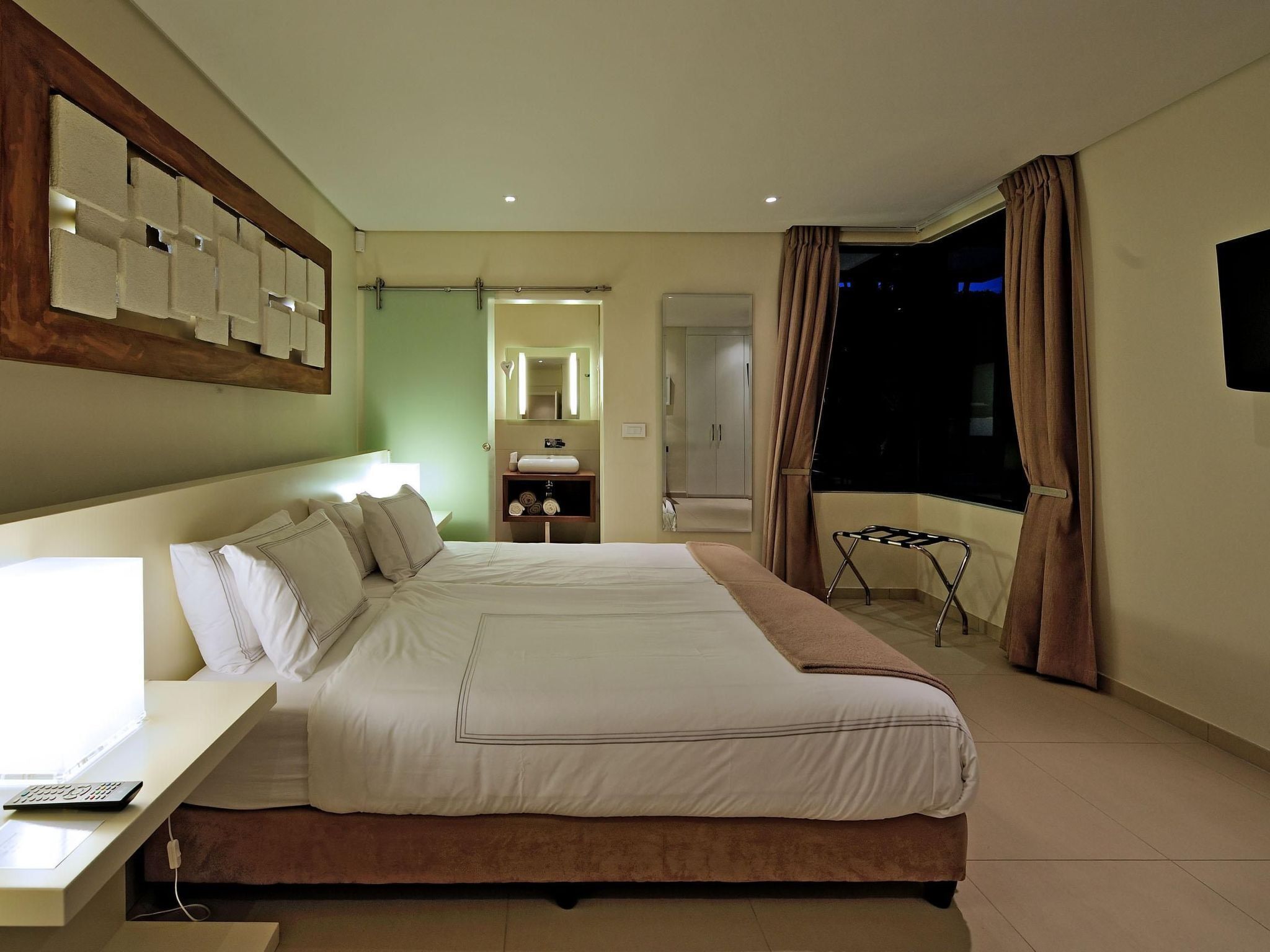 Een hotelkamer met een groot bed, een bureau met een computer, een spiegel, een wastafel met een wasbak en uitzicht op de nachthemel door het raam.