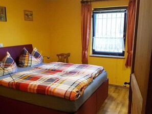 Een slaapkamer met een bed, een houten stoel, een raam met gordijnen en een muur met twee afbeeldingen.