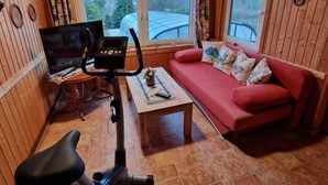 L’intérieur d’un chalet en bois est aménagé avec un canapé rouge, un tapis de course, un téléviseur et de grandes fenêtres qui offrent une vue sur les arbres. Le toit est incurvé.