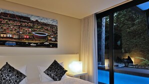 Moderni hotellihuone, jossa on suuri sänky, kaupunkimaisemaa esittävä taulu, näkymä yölliseen kaupunkiin ikkunasta sekä pieni pöytä, jossa on lamppu.