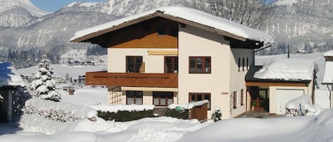 Un chalet de dos pisos con balcón de madera, rodeado de montañas nevadas.
