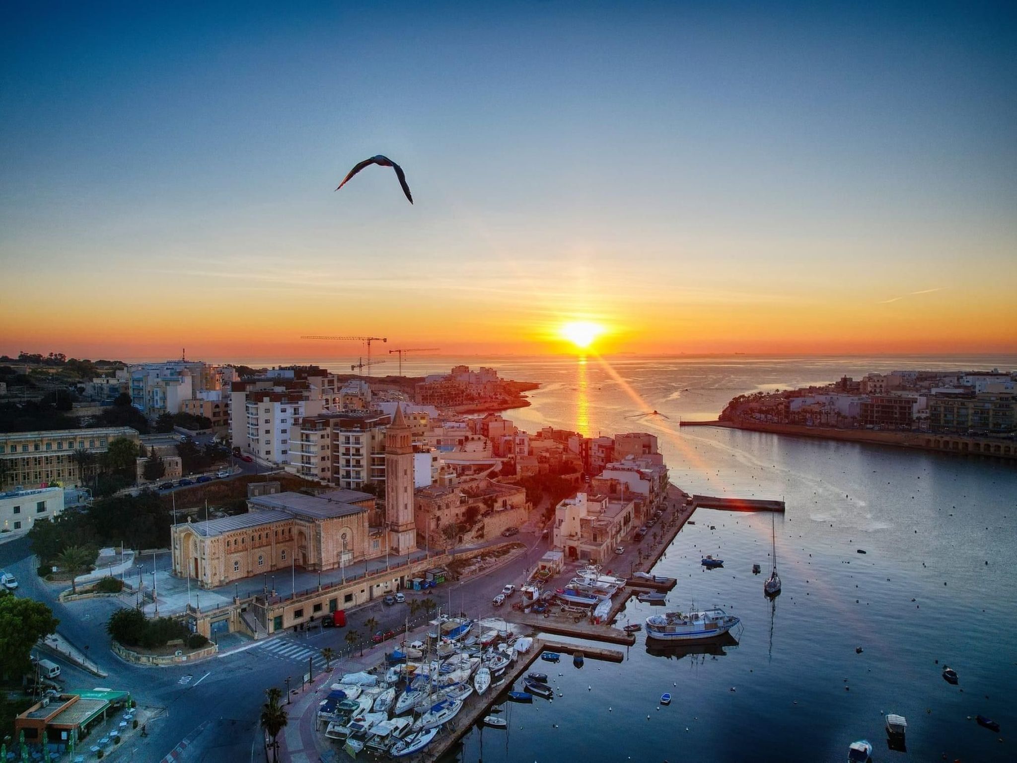 En kystby ved solnedgang med en markant historisk bygning, en havn med både og et klart syn på solnedgangen over vandet.