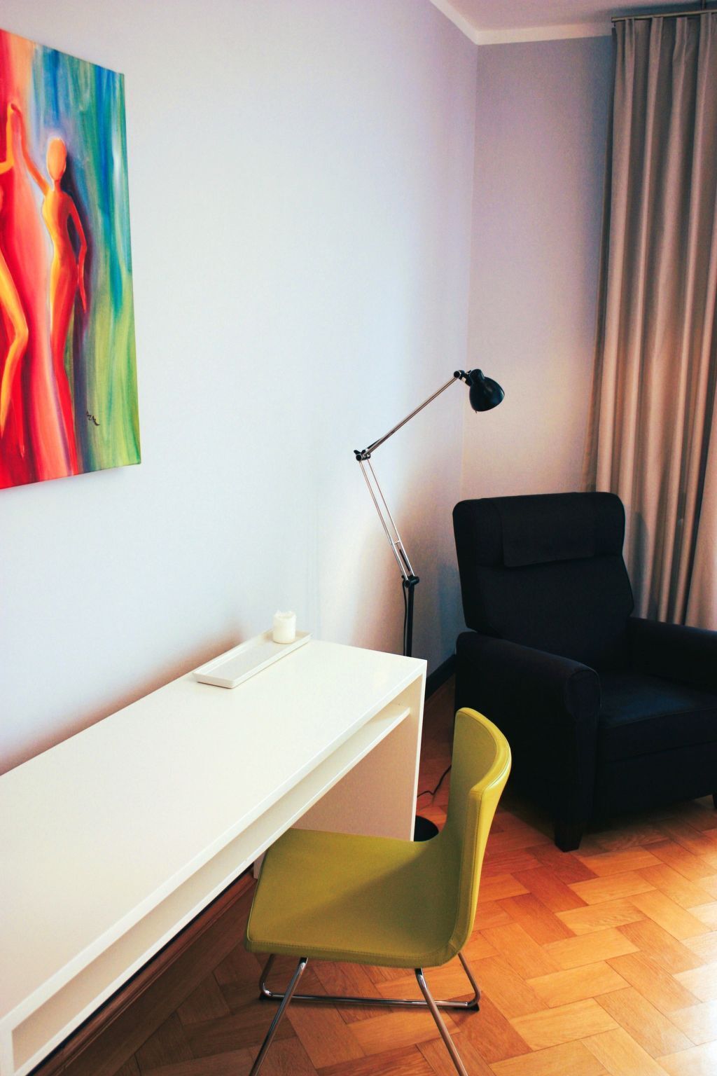 Une pièce avec un bureau blanc, une chaise noire, une chaise verte, un lampadaire et un tableau accroché au mur.