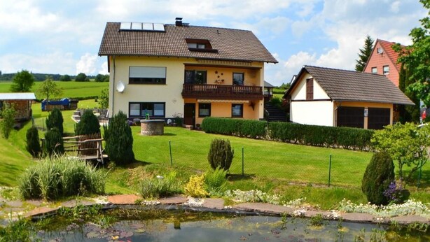 Une maison à deux étages avec un balcon, un jardin agrémenté d’un bassin, et un bâtiment à toit rouge en arrière-plan.