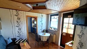 Un salon chaleureux avec un plafond en bois, un coin repas avec une petite table et des chaises, et une cuisine à l’arrière-plan.