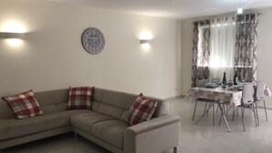 A living room with a beige sectional sofa, two plaid cushions, a wall clock, and a dining area with a table set for two. - 3. Appartement mit Meerblick in Strandnähe by Interhome (Marsascala)