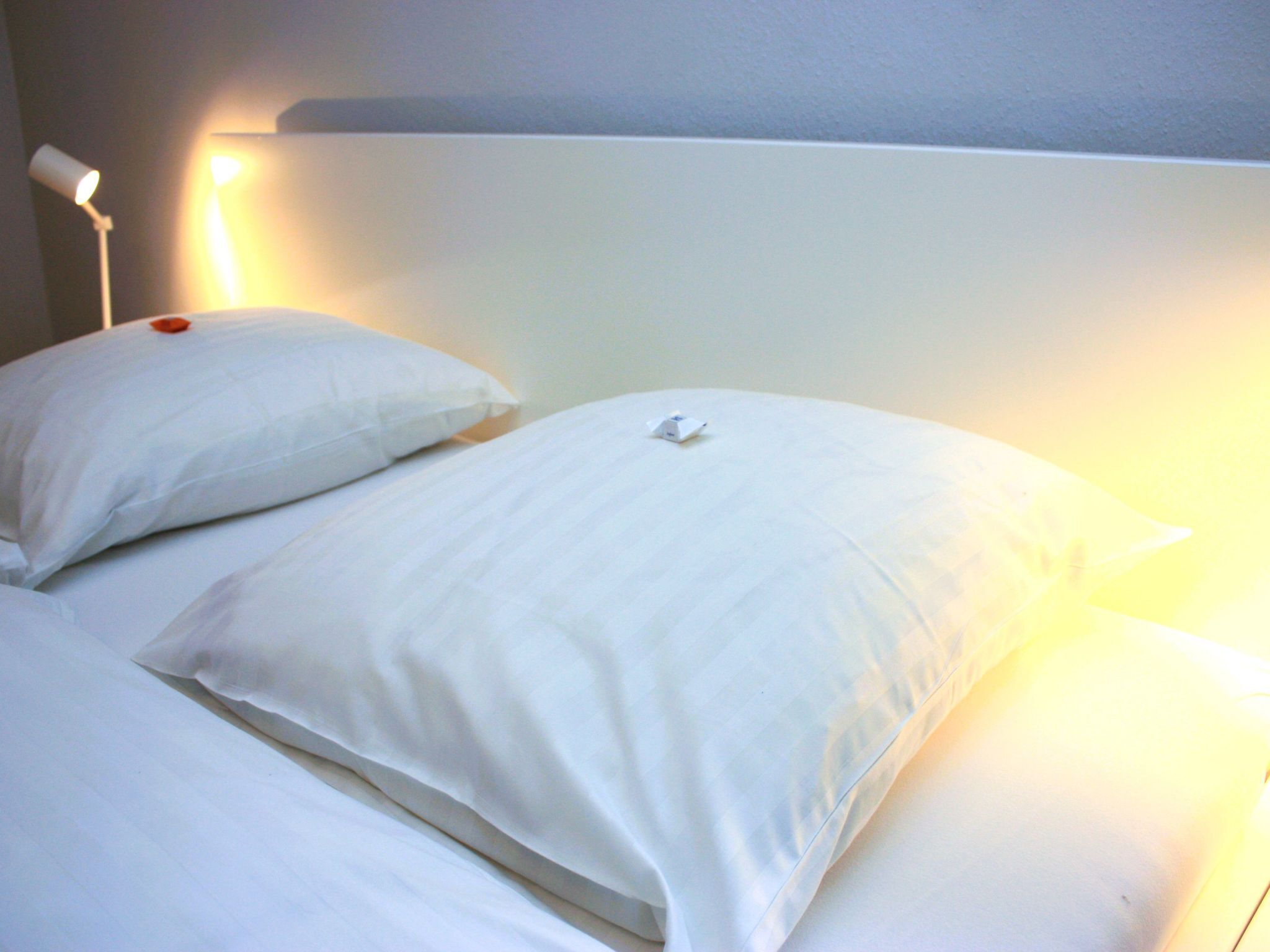 Ein ordentlich bezogenes Bett mit weißen Leinen und Kissen, ein weißer Kopfteil und eine Nachttischlampe, die sanftes Licht spendet.